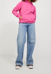 Kuumansävyinen vaaleanpunainen huppari kengurun taskulla, yhdistetty vaaleansinisiin leveälahkeisiin farkkuihin ja mustiin tennareihin. Puuvillainen materiaali, rento muotoilu.