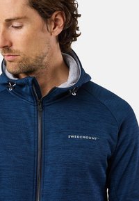 Giacca blu con zip realizzata in tessuto testurizzato. Presenta un cappuccio interno grigio, una zip frontale e un accento con logo. Design aderente.