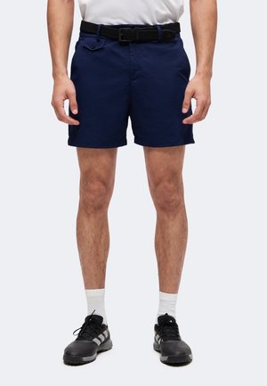 Man draagt marineblauwe korte broek met zwarte riem, witte sokken en zwarte sportschoenen, staand tegen een effen achtergrond.