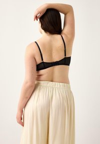 Reggiseno in rete nera con spalline regolabili e chiusura a gancio, abbinato a pantaloni fluidi color crema, che mettono in mostra una texture liscia.
