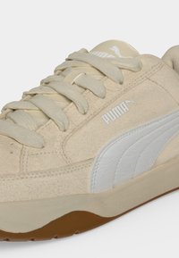 Μπέζ σουέντ αθλητικό παπούτσι Puma με λευκό λογότυπο και ρίγα, μπεζ κορδόνια και λαστιχένια σόλα, εμφανίζεται σε κοντινή πλευρική όψη.