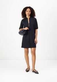 Navyblaues Kurzarmkleid mit Reißverschluss am Hals und zwei vorderen Taschen. Das Model hält eine schwarze, geraffte Handtasche und trägt schwarze flache Schuhe.