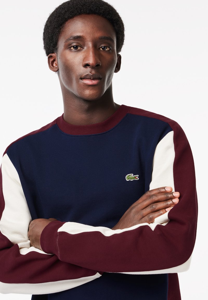Sudadera azul marino con mangas en color burdeos y blanco, que presenta un pequeño logotipo de cocodrilo verde. El material parece suave y texturizado.