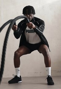 Fekete sportdzseki fehér díszítéssel egy szürke póló fölött, fekete rövidnadrág és fekete sportcipő. Egy vastag, texturált harci kötelet fog.