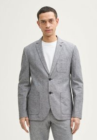 Grau strukturierter Blazer mit zwei vorderen Taschen und Reverskragen, ausgestattet mit zwei schwarzen Knöpfen und einer einzelnen Brusttasche. Kombiniert mit einem weißen T-Shirt.