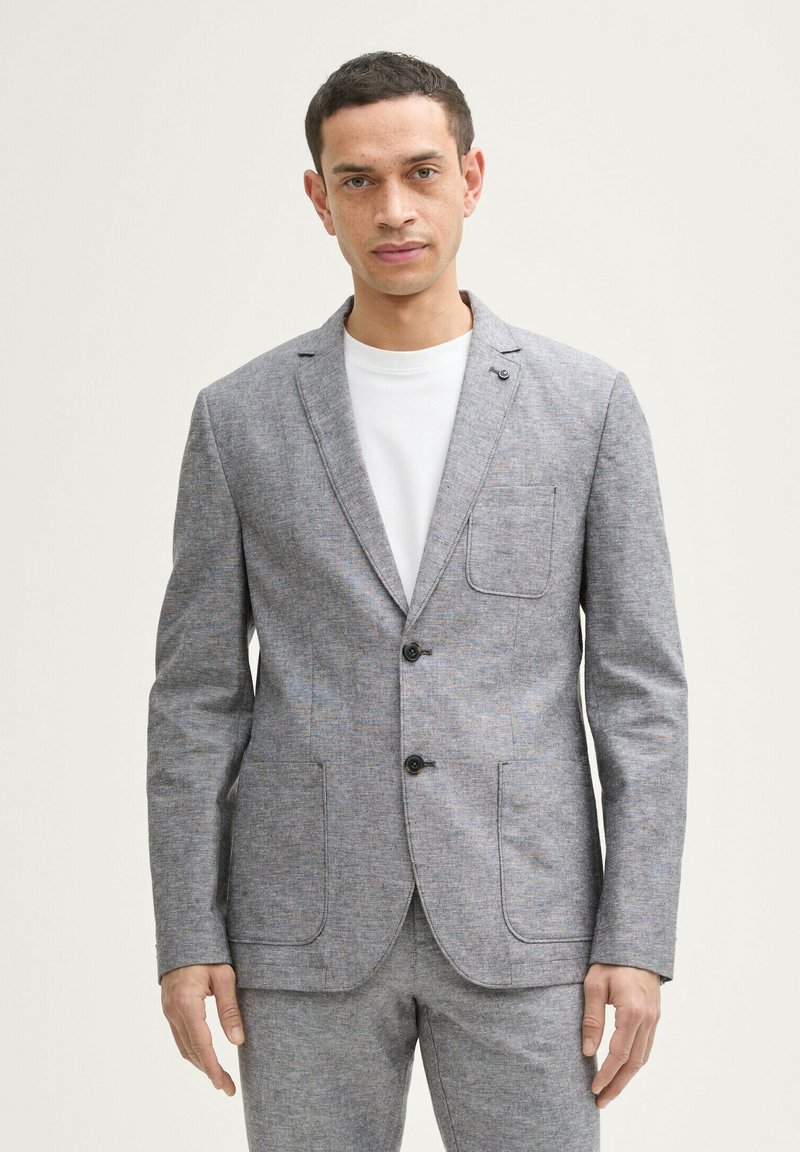 Grau strukturierter Blazer mit zwei vorderen Taschen und Reverskragen, ausgestattet mit zwei schwarzen Knöpfen und einer einzelnen Brusttasche. Kombiniert mit einem weißen T-Shirt.
