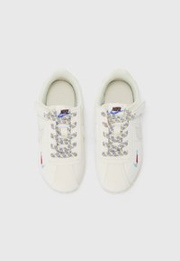 Par en hvite Nike-sneakers med mønstrete lisser og små fargerike Nike swoosh-brodere på sidene, sett ovenfra på hvit bakgrunn.