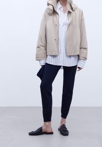 Persona che indossa una giacca beige, una camicia a righe bianche, pantaloni jogger blu navy e scarpe slip-on nere, in piedi su un pavimento grigio contro un muro bianco.