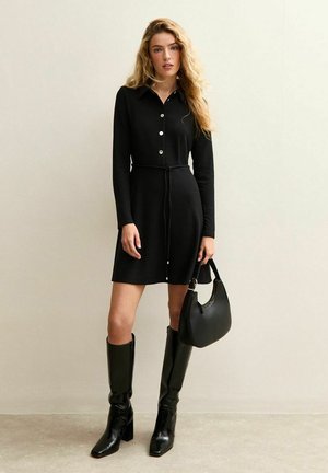 Donna con capelli biondi ricci che indossa un vestito nero a maniche lunghe abbottonato, tiene in mano una borsa nera e indossa stivali neri al ginocchio.