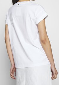 Camisa de algodón blanca con mangas enrolladas, que presenta un pequeño logotipo en la parte posterior del cuello y un dobladillo recto, vista desde atrás.