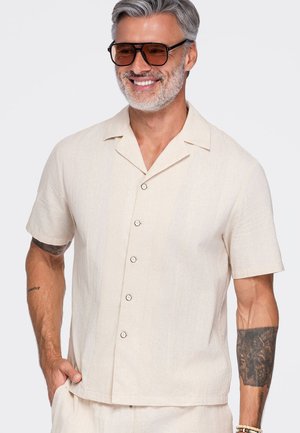 Homme aux cheveux gris et à la barbe, portant des lunettes de soleil marron et une chemise beige clair à manches courtes boutonnée, souriant avec une main dans la poche.