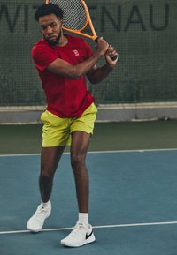 En mannlig tennisspiller svinger en racket, iført en rød skjorte og lysegule shorts, supplert med hvite og svarte joggesko.