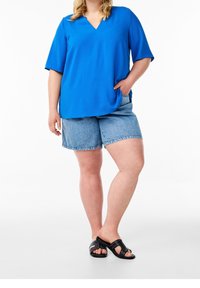 Sininen V-kaula-aukkoinen paita lyhyillä hihoilla yhdistettynä vaaleansinisiin farkkushortseihin. Mallilla on päällään mustat liukusandaalit, joissa on niittikoristelu.