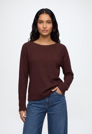Femme aux longs cheveux foncés portant une chemise marron à manches longues et un jean bleu, debout avec une main dans la poche devant un fond uni.