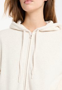 Sweat à capuche beige clair avec fermeture éclair, en tissu doux et texturé, doté de cordons de serrage et d'une capuche. La fermeture éclair est métallique et le design est minimaliste.