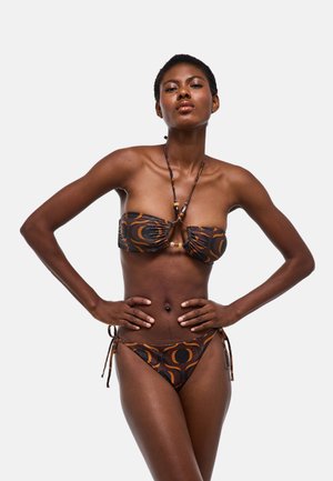 Femme en bikini bandeau marron et noir à motif, les mains sur les hanches sur un fond blanc uni.