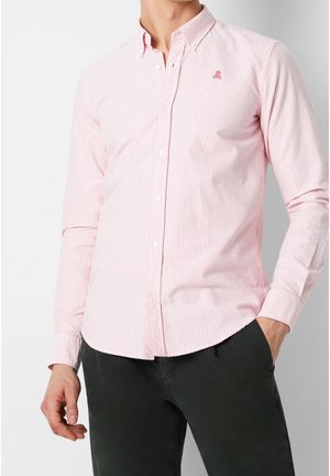 Hombre vistiendo una camisa entallada de botones con rayas rosas y blancas, con un pequeño logo de calavera en el pecho, combinado con pantalones oscuros.