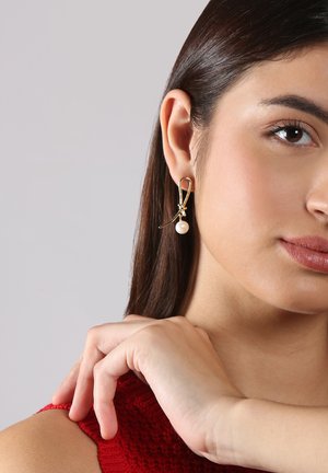 Goldfarbene Ohrringe mit einem Schleifen-Design, ausgestattet mit einem kleinen Perlenanhänger, vor einem neutralen Hintergrund.