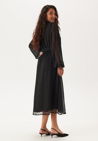 Svart midikjol med prickig struktur, långa transparenta ärmar och knytmidja. Bärs med svarta spetsiga klackskor. Slät tyg.