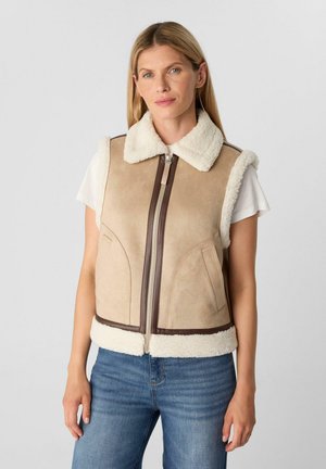 Kobieta w beżowej kamizelce z kożuszkiem, zapinanej na zamek i z kieszeniami, na białej koszulce z krótkim rękawem i niebieskich dżinsach, stojąca na jednolitym tle.