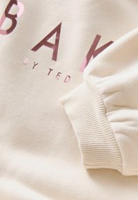 Rękaw beżowego swetra z prążkowanym mankietem obok częściowego napisu "BAK BY TED" dużymi, błyszczącymi różowymi i brązowymi literami.