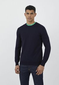 Maglione blu navy a maglia con scollatura rotonda, maniche lunghe e polsini a coste. Indossato sopra una maglietta verde, abbinato a jeans scuri.