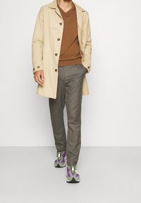Beige trench coat, hnědý svetr s véčkovým výstřihem, vzorované šedé kalhoty a multikulturní tenisky s akcenty v zelené, fialové a šedé barvě.