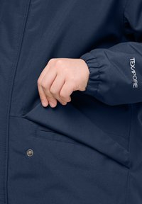 Námořnická modrá nepromokavá bunda s hladkou texturou, vybavená elastickým manžetovým zakončením a bočním kapsou se stříbrným zacvakávacím uzávěrem.