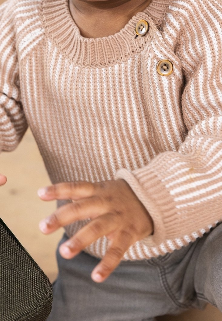 Tout-petit portant un pull en tricot beige avec des boutons à l'épaule et un pantalon gris, tendant les deux mains vers l'avant.