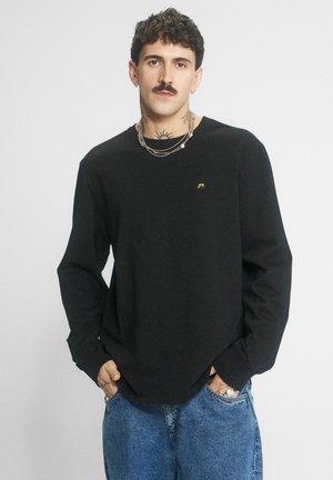 Mężczyzna z wąsami, ubrany w czarną koszulę z długim rękawem, warstwowe łańcuchy i niebieskie dżinsy, stoi z rękami w kieszeniach na jednolitym tle.