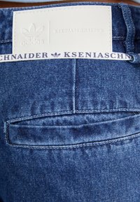 Denim kavbojke z svetlo modrim pranjem, vidno šivano, s hrbnimi žepi in oznako z blagovno znamko v belih in mornarsko modrih odtenkih.