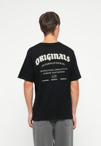 Juodas medvilnės marškinėliai, kuriuose ant nugaros didelėmis baltomis raidėmis užrašyta "ORIGINALS INTERNATIONAL" ir papildomas tekstas mažesniu šriftu.