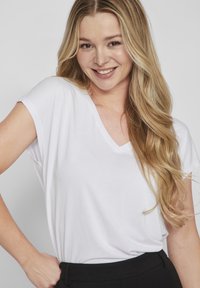 Biała koszulka typu V-neck z luźnymi rękawami, miękkiego materiału i luźnym fasonem. Modelka pozuje z ręką na biodrze, nosząc czarne spodnie.