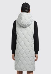 Hellgraue, gesteppte Pufferweste mit Kapuze, die ein Rautenmuster aufweist und darunter ein schwarzes Langarmshirt trägt. Das Markenlogo ist sichtbar.