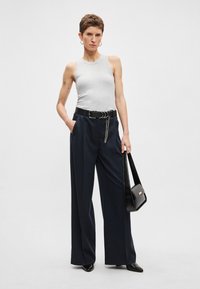 Szürke ujjatlan felső, tengerészkék bő szárú nadrág, fekete öv láncos díszítéssel és fekete válltáska. Sima anyagok; minimalista dizájn.