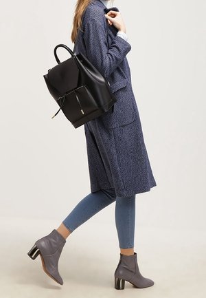 Femme en manteau bleu et jean marchant, portant un sac à dos en cuir noir et des bottines à talons grises.