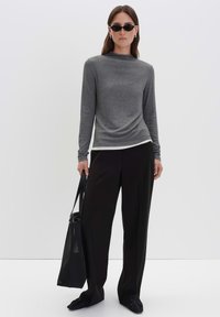 Frau mit schwarzen Sonnenbrillen, grauem Langarmoberteil, weiten schwarzen Hosen, schwarzen Schuhen, die eine große schwarze Netztragetasche vor einem weißen Hintergrund hält.