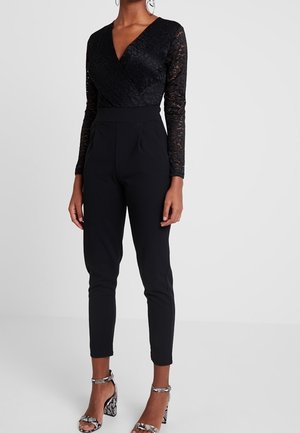 Femme portant un haut en dentelle noire à manches longues avec un col en V, un pantalon noir ajusté taille haute et des escarpins à motif peau de serpent, debout sur un sol gris.