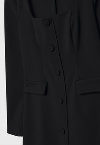 Manteau noir cintré avec boutons recouverts sur le devant et deux poches à rabat au niveau de la taille, vue rapprochée.