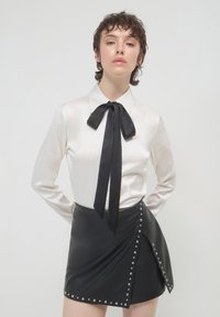 Giovane con capelli corti e ricci indossa una camicetta bianca con cravatta nera a fiocco e una gonna di pelle nera borchiata, in posa con le mani dietro la schiena.