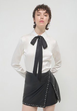 Young person with short curly hair wears white blouse with black bow tie and black studded leather skirt, posing with hands behind back.