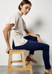 Hellbeige strukturierte Kurzarmbluse mit einem hohen Kragen, kombiniert mit dunkelblauen Jeans und braunen Slippern, sitzt auf einem Holzschemel.