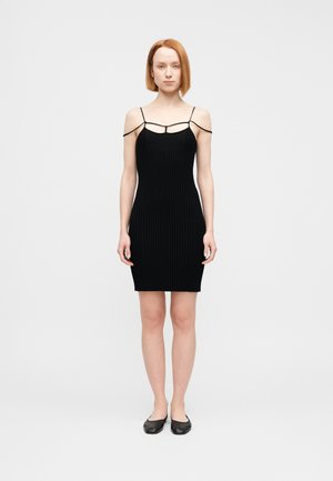 Czarna prążkowana sukienka na cienkich ramiączkach, dopasowany krój, długość do połowy uda, falowany dół. Modelka nosi czarne baleriny.