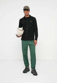Camicia nera a bottone, pantaloni verdi, scarpe da ginnastica grigie, con una borsa di colore crema, indossando un cappellino a scacchi marrone. Sfondo semplice.