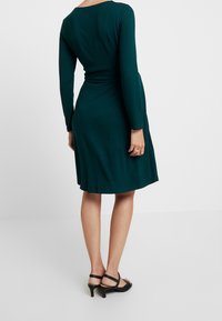 Mörk teal långärmad klänning med figursydd midja och A-linjeformad kjol. Slätt tyg med rund halsringning, i kombination med svarta klacksandaler.