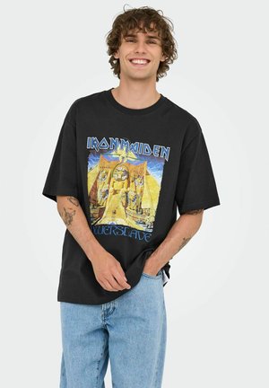 Camiseta de algodón negra con un diseño gráfico de un faraón y pirámides, con el texto "IRON MAIDEN POWERSLAVE" en azul y amarillo. Corte relajado.