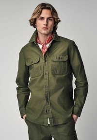 Olive green button-up shirt made of soft fabric, featuring two chest pockets and brown buttons, worn over a white shirt with a red scarf.