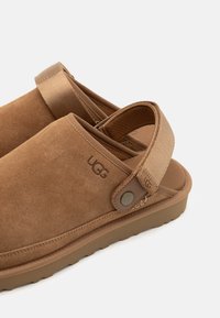 Brown suede slip-on shoes with a textured strap, featuring UGG branding and a rubber sole. The design includes a cushioned footbed and open back.