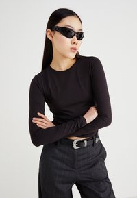 Bluză neagră cu mâneci lungi și design ajustat; purtată cu pantaloni gri cu dungi și o curea cu cataramă argintie; accesorizată cu ochelari de soare negri cu ramă.