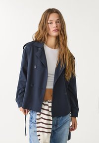 Mörkblå trenchcoat med dubbelknäppt design, texturerad tyg, axelremmar och två sidofickor. Matchas med en vit topp och randig tröja.